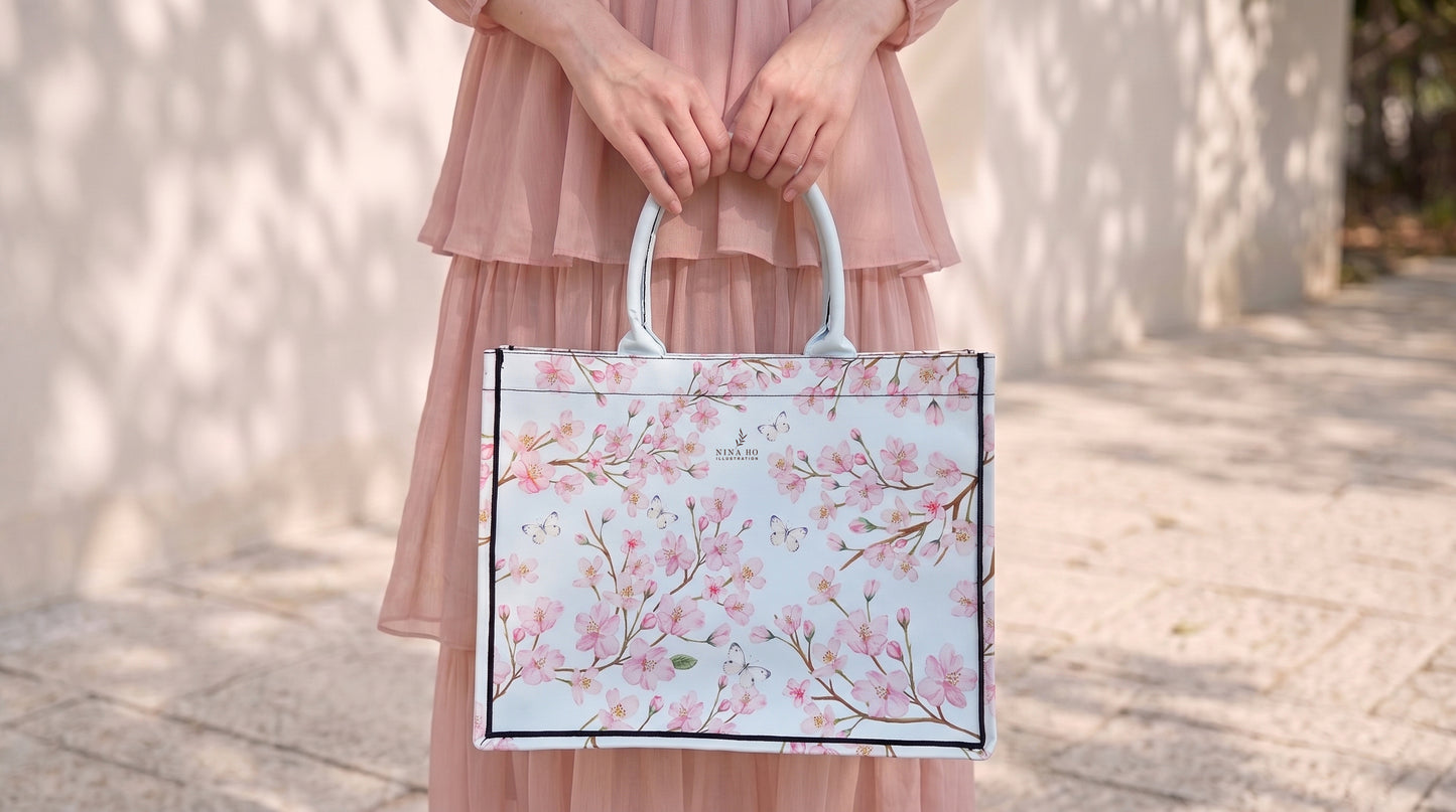 A woman in a soft pink dress holds a structured canvas tote bag adorned with delicate hand-painted cherry blossom branches, embodying subtle botanical elegance.