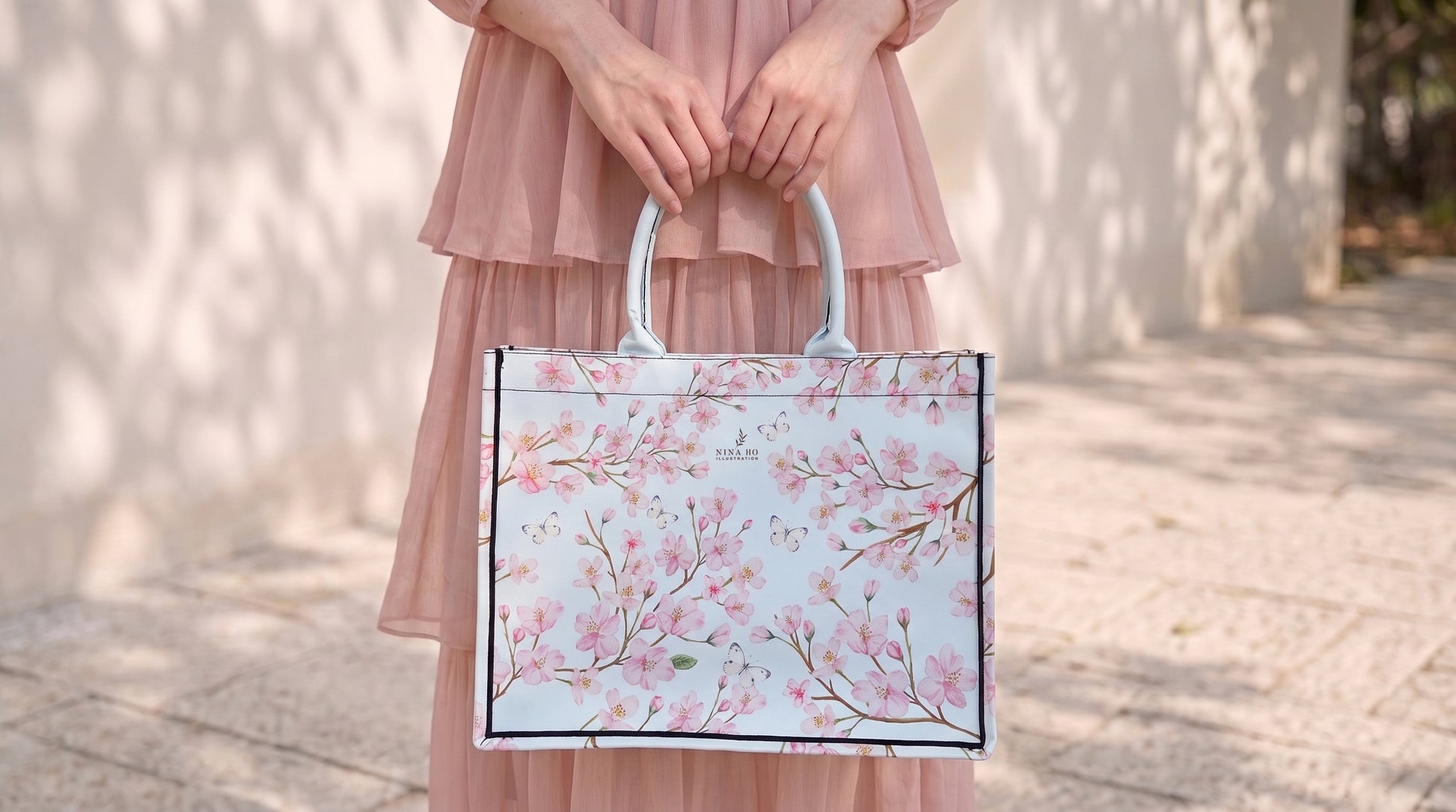 A woman in a soft pink dress holds a structured canvas tote bag adorned with delicate hand-painted cherry blossom branches, embodying subtle botanical elegance.