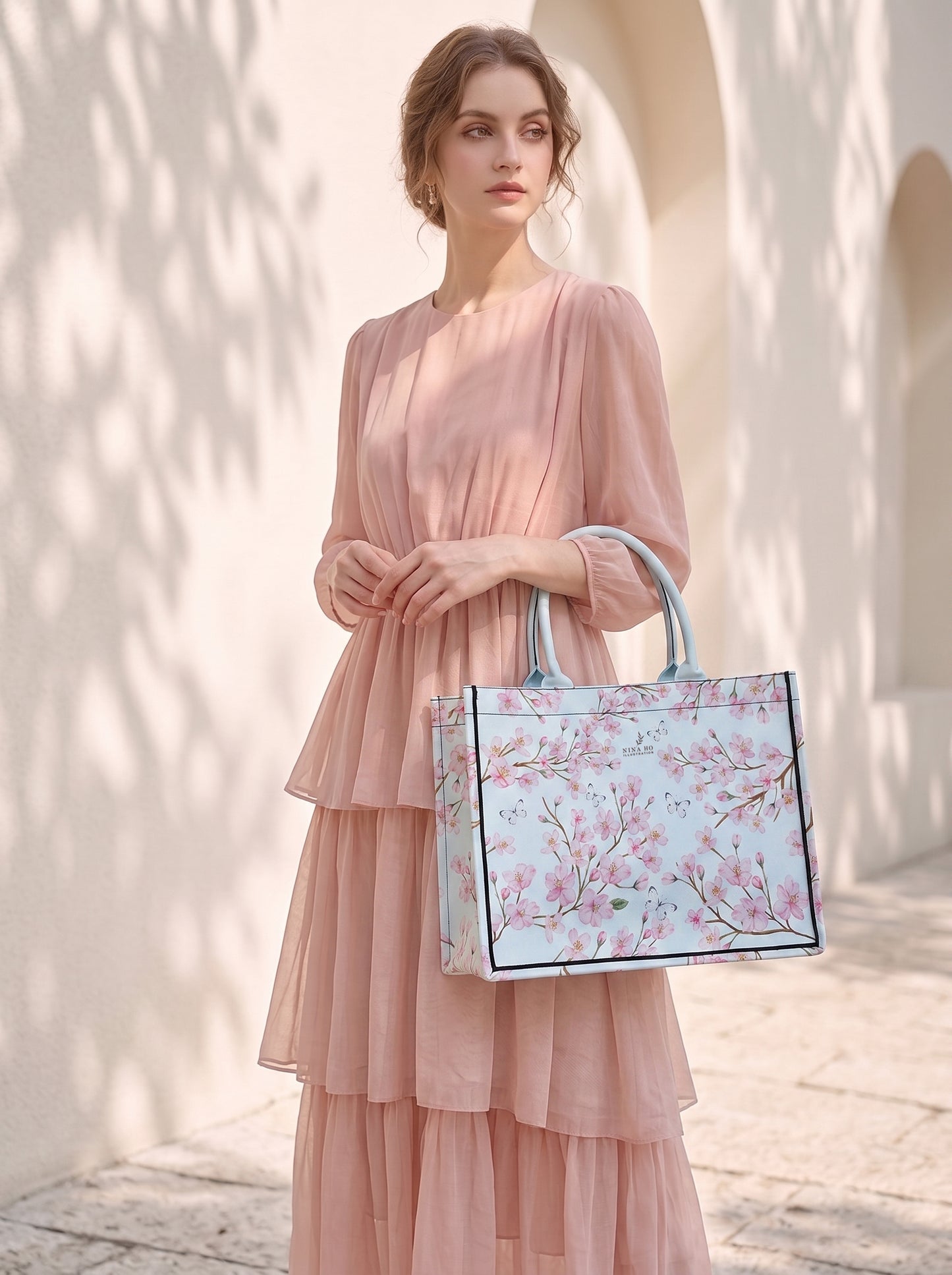 A woman in a soft pink, tiered chiffon dress stands gracefully against an arched, sunlit wall, holding a structured tote adorned with delicate cherry blossom branches and pale pink flowers, evoking refined botanical elegance.