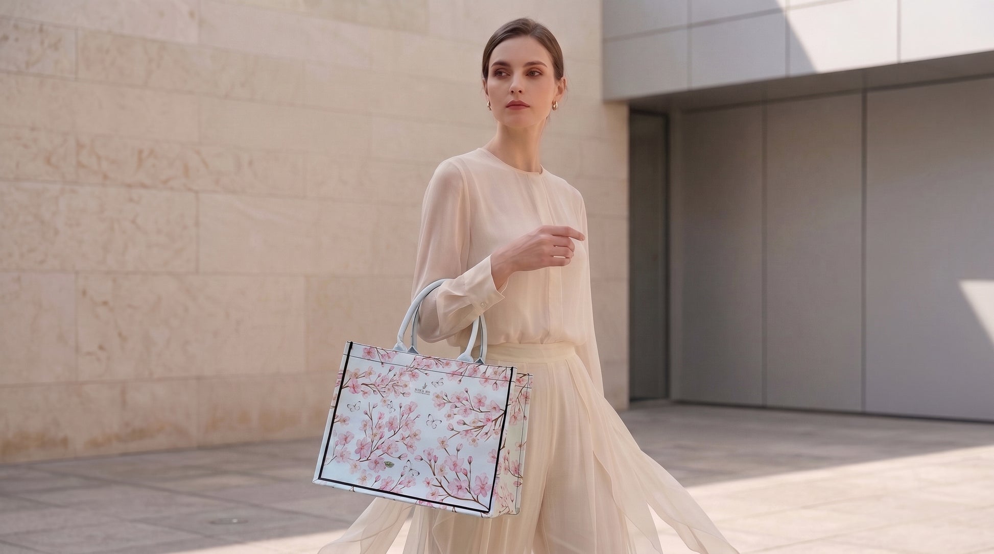Elegant woman in a flowing cream dress carries a pale blue canvas tote adorned with hand-painted cherry blossom motifs by NINA HO ILLUSTRATION, blending artistic subtlety with refined everyday sophistication.