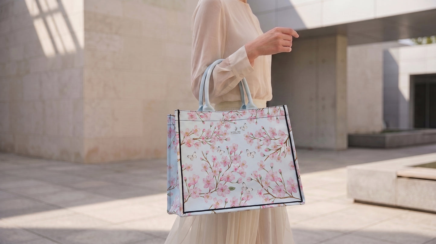 A sophisticated canvas tote in pale blue, adorned with delicate hand-painted cherry blossom branches and fluttering white butterflies, reflecting NINA HO ILLUSTRATION’s refined botanical artistry.