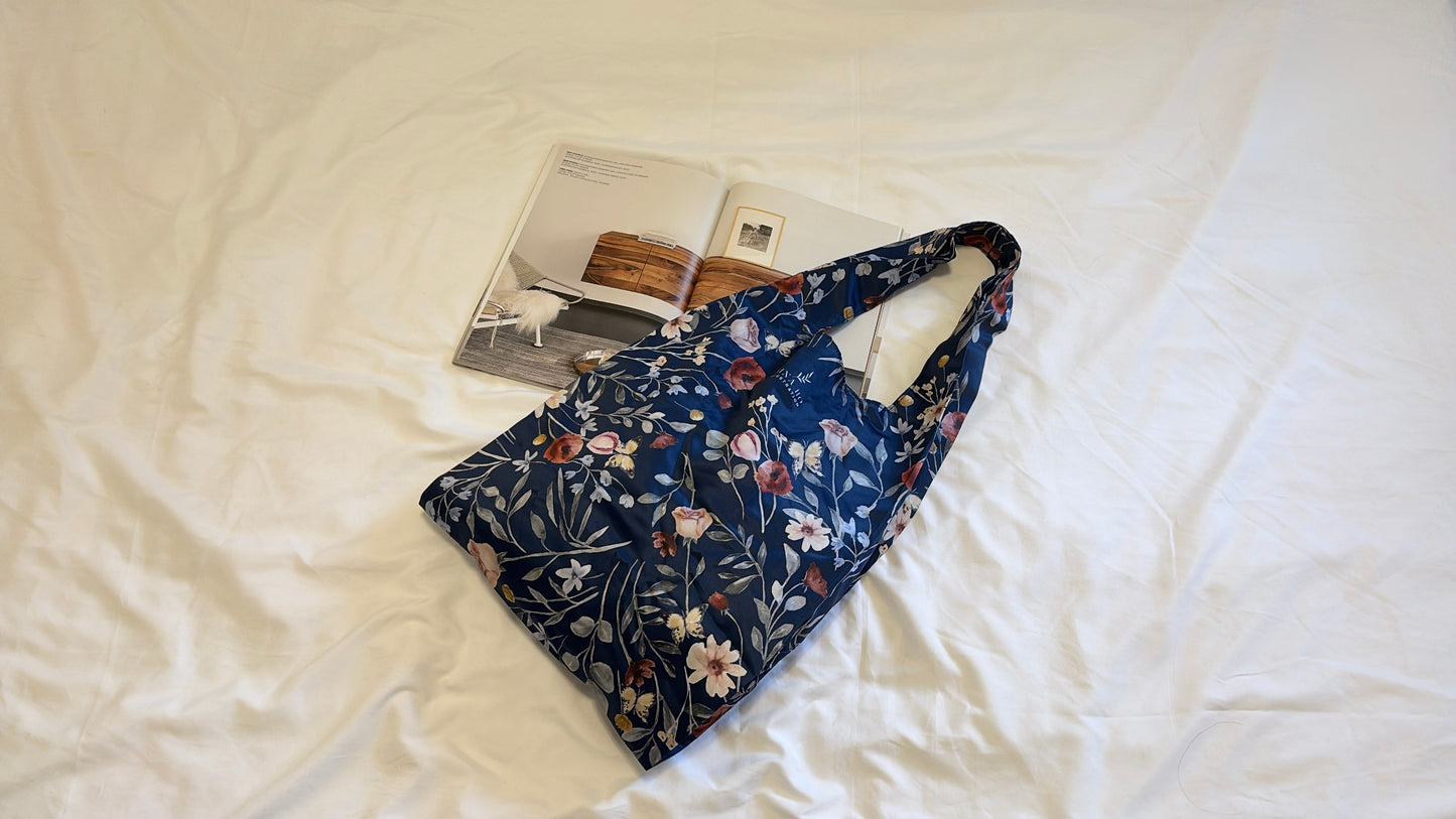 A dark blue foldable tote with delicate hand-painted floral motifs featuring small pink and white blossoms, elegantly resting on a crisp white fabric backdrop beside an open book.