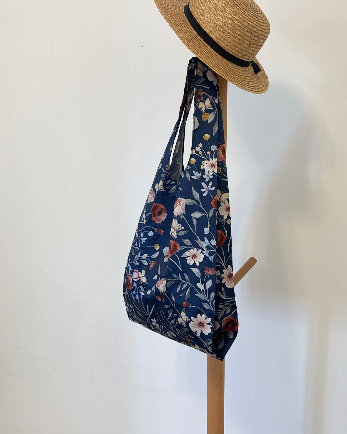 A dark blue foldable tote bag adorned with a delicate botanical pattern of white and deep red flowers and slender green leaves, hanging alongside a woven straw hat with a black ribbon.