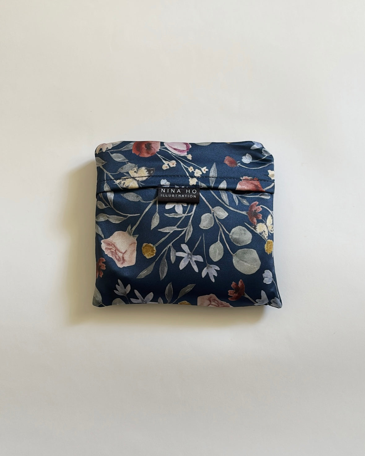 A neatly folded dark blue silk accessory adorned with delicate hand-painted botanical illustrations featuring pale pink blossoms, white star-shaped flowers, and soft green leaves, embodying refined Parisian chic.