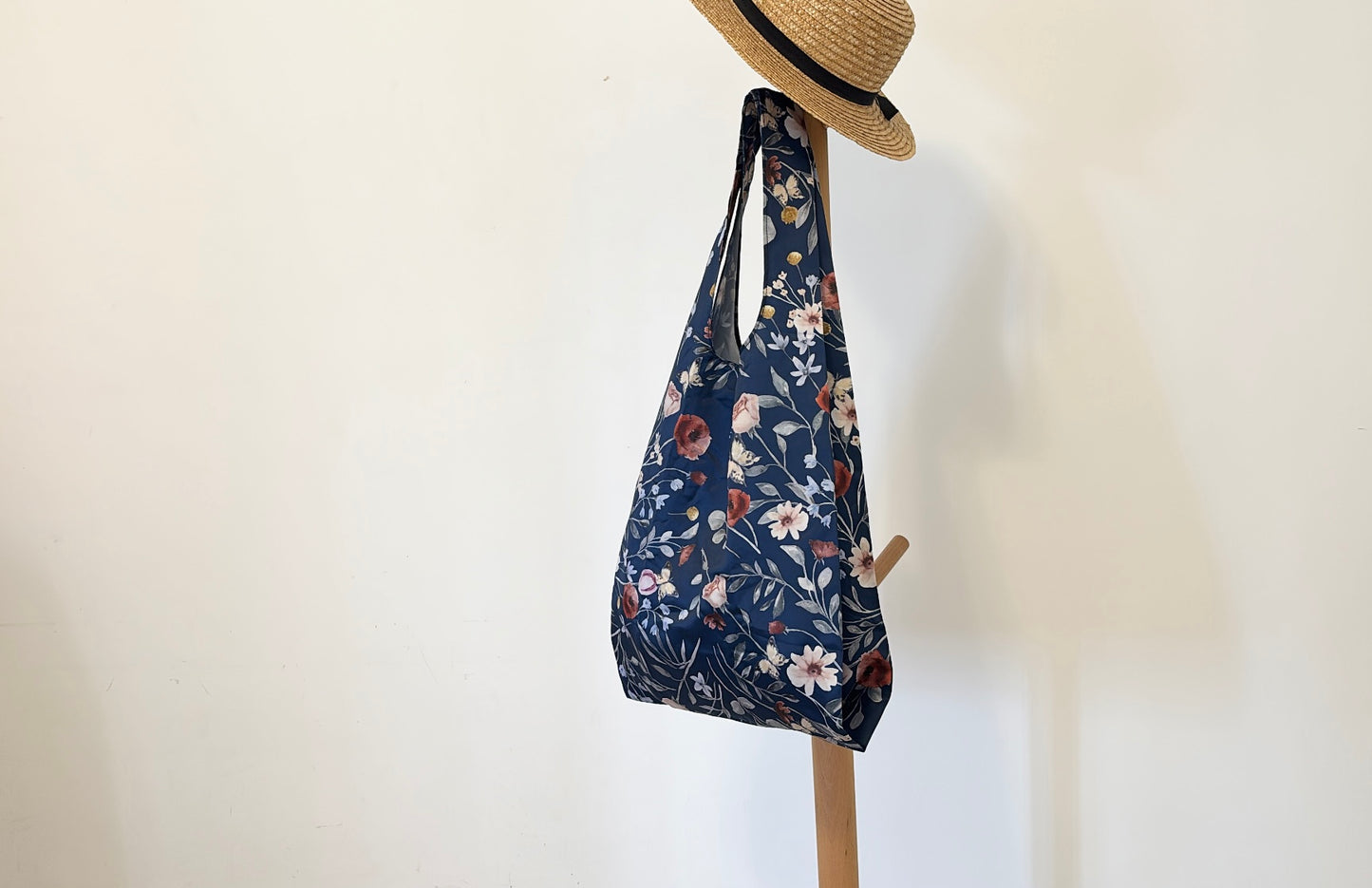 Dark blue botanical foldable tote bag adorned with delicate floral patterns, suspended beneath a classic straw boater hat on a minimalist wooden stand.