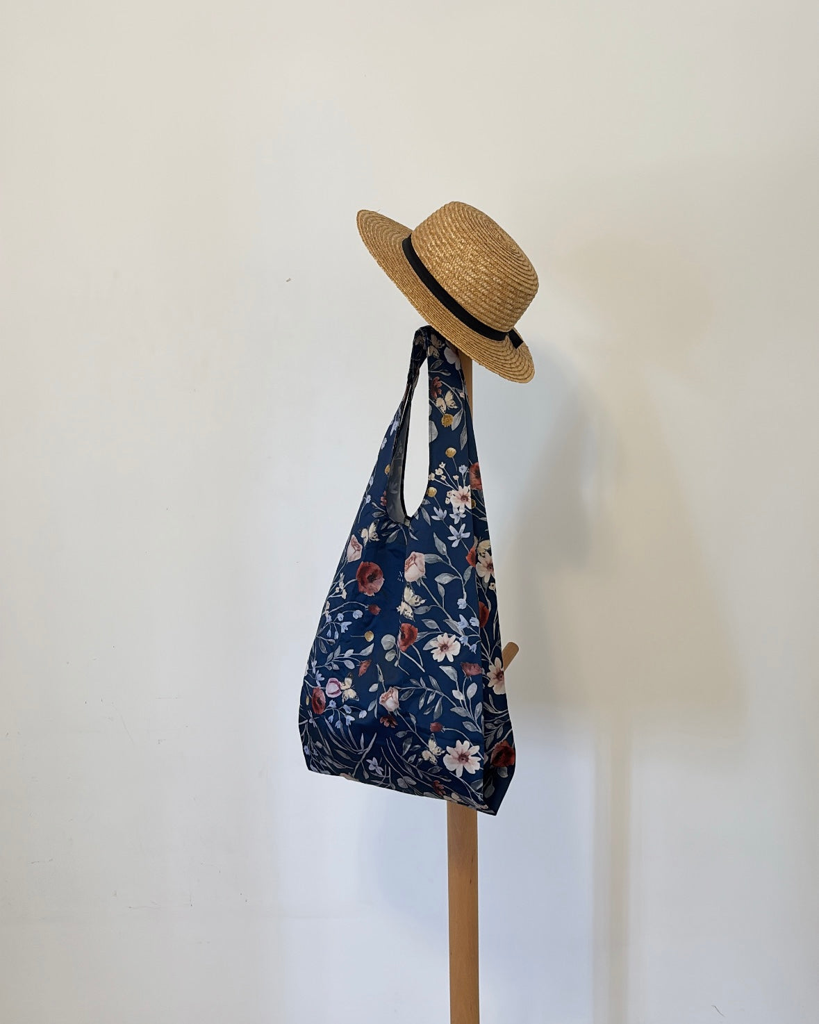 A deep blue botanical print foldable tote adorned with delicate white and muted red flowers, suspended beneath a classic straw hat, evoking refined Parisian chic and curated elegance by NINA HO ILLUSTRATION.