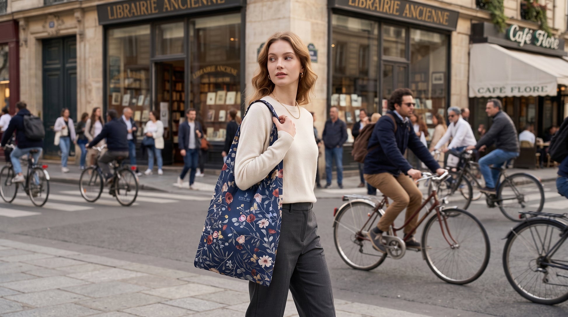 A woman in tailored dark trousers and a cream top carries a dark blue botanical print tote bag on a bustling Parisian street, embodying understated urban elegance.