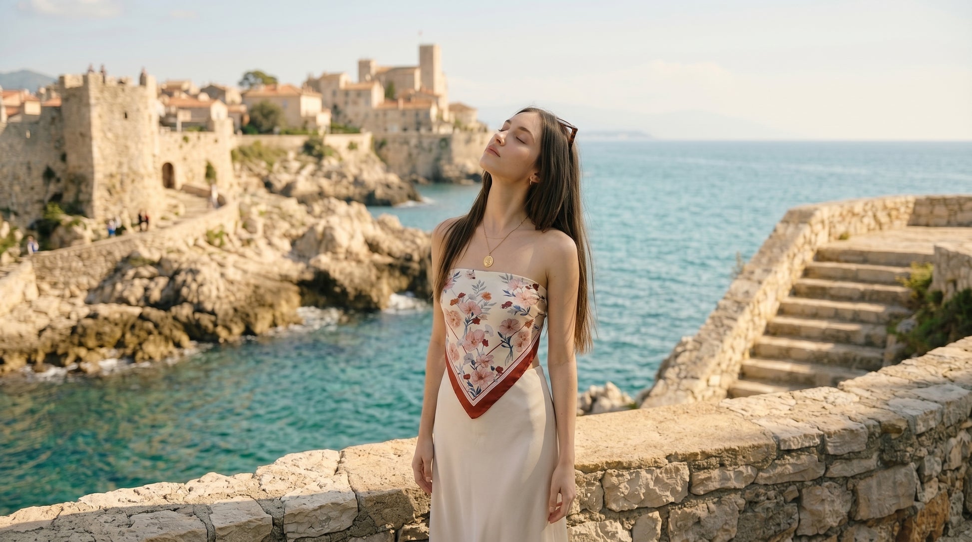 A woman exudes effortless Parisian chic wearing a NINA HO ILLUSTRATION double-sided 100% mulberry silk scarf, adorned with hand-painted Icelandic poppies and delicate chocolate cosmos in warm orange-red and soft blue-purple tones, elevating her elegant ivory dress against a sunlit coastal backdrop.