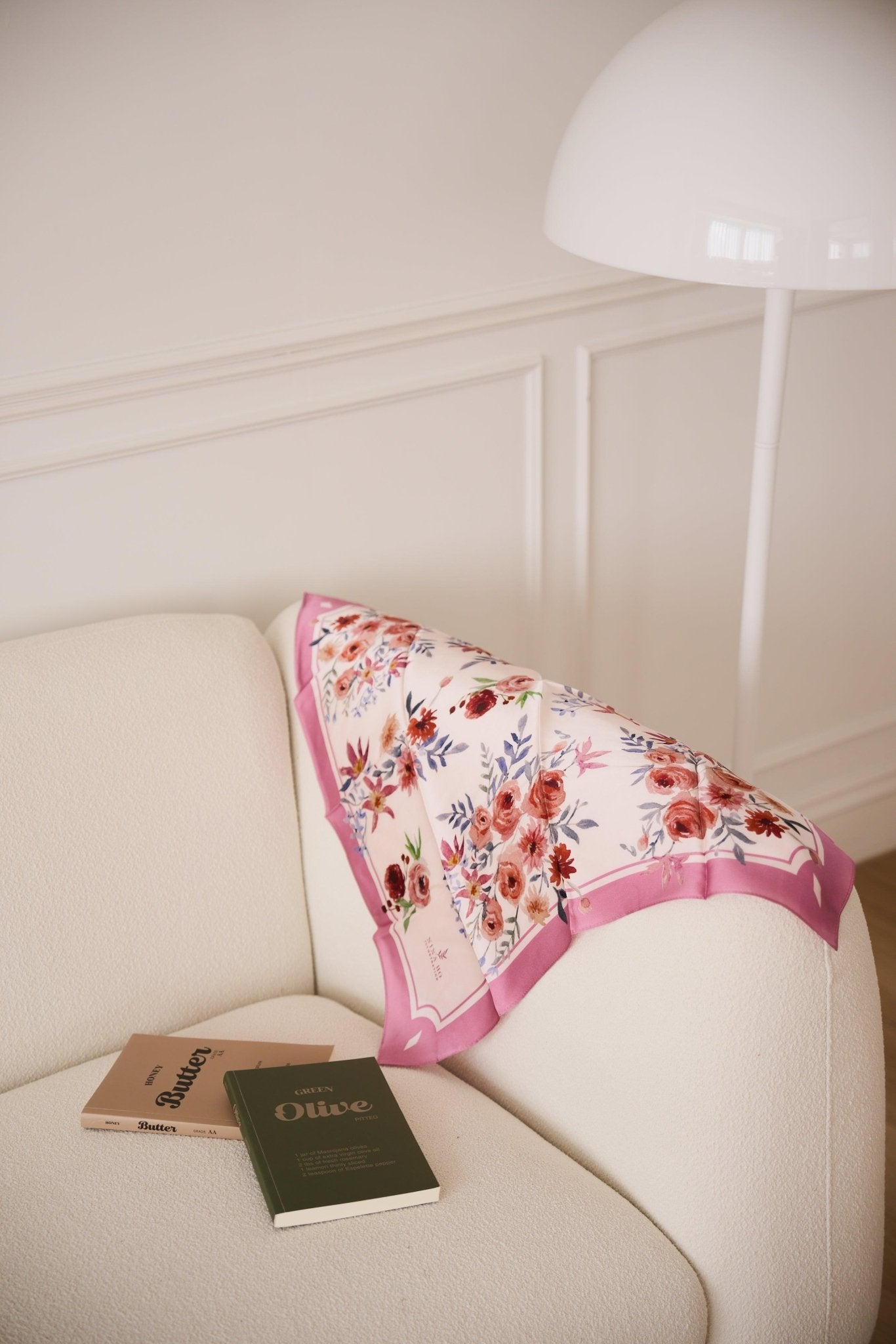 Delicate 100% mulberry silk scarf draped on cream sofa, adorned with hand-painted ranunculus and purple clematis, embodying Parisian chic by Nina Ho Illustration.